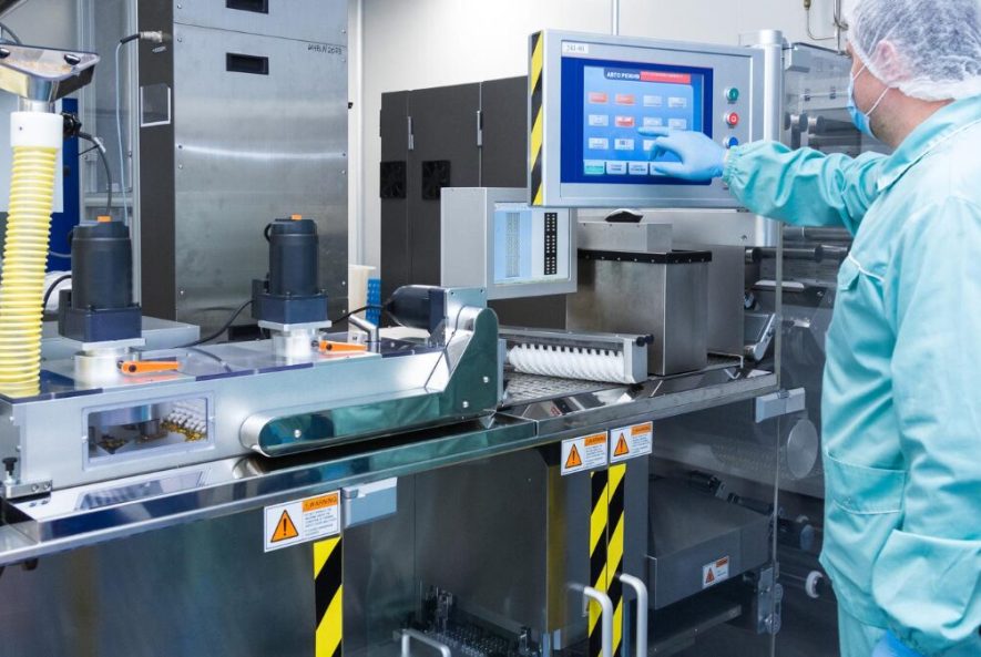 Pharmaceutical factory worker in sterile protective clothing operating automated blister packaging machinery using a digital touch screen.